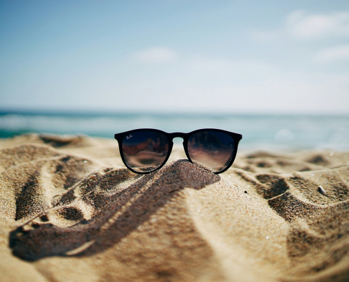 Eine Sonnenbrille steckt halb in einem Sandhügel am Strand, mit dem Meer und einem blauen Himmel im Hintergrund.