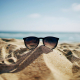 Eine Sonnenbrille steckt halb in einem Sandhügel am Strand, mit dem Meer und einem blauen Himmel im Hintergrund.