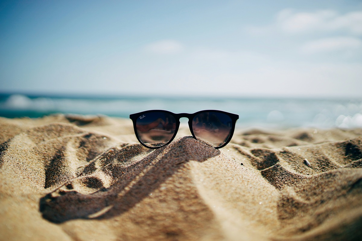 Eine Sonnenbrille steckt halb in einem Sandhügel am Strand, mit dem Meer und einem blauen Himmel im Hintergrund.